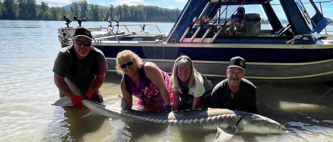 A group of four people is standing in shallow water, smiling at the camera while holding a large sturgeon. Behind them, there is a blue and white boat equipped with fishing rods, and a forested shoreline is visible in the background under a clear sky. - 7 Safety Tips for Ensuring a Safe and Enjoyable Fishing Experience in the Summer Heat