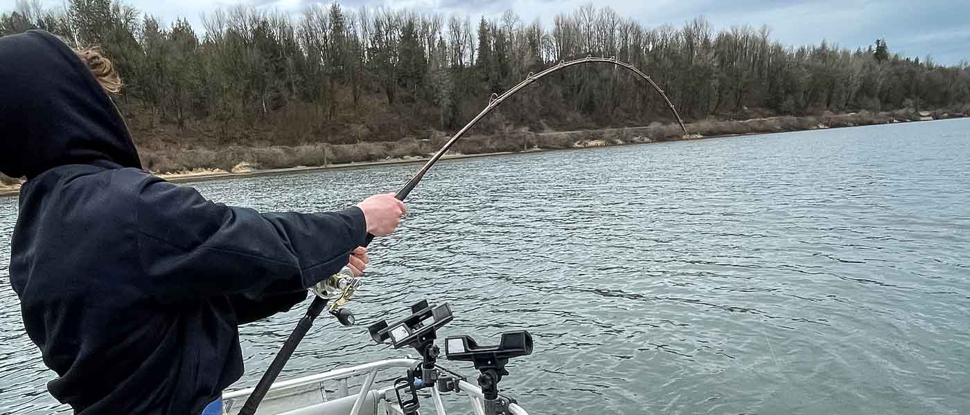 Man in a hoodie attempts to reel in a fish off the rear of a boat.